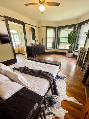 Charming bedroom with large bay windows, cozy decor, and modern accents, featuring a plush bed and vibrant indoor plant.