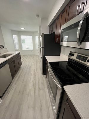 Modern kitchen with sleek stainless steel appliances, dark wood cabinetry, and stylish light-colored flooring leading to a bright dining area with large windows.