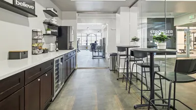 Modern communal area featuring sleek countertops, stylish bar seating, and motivational decor, leading to a glass-enclosed gym space.