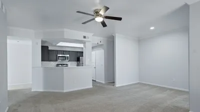Bright and airy living area with plush carpeting, ceiling fan, and open-access kitchen showcasing modern stainless steel appliances.