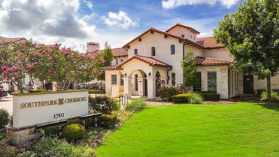 Charming Mediterranean-style apartment complex with lush landscaping and colorful flowering trees under a bright blue sky.