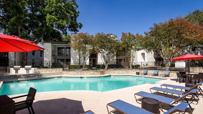 Inviting apartment complex with a sparkling pool, ample lounge chairs, and vibrant red umbrellas, perfect for relaxation and enjoying sunny weather.