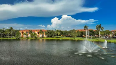 Scenic lakeside view of elegant apartment buildings with lush palm trees and a vibrant fountain under a clear blue sky.