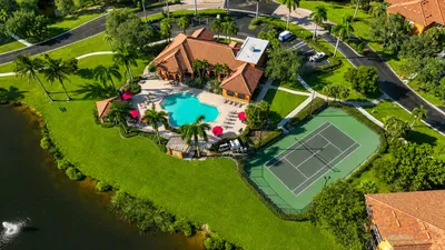 Aerial view of luxurious apartment complex featuring a resort-style pool with lounge chairs and vibrant umbrellas, adjacent to a well-maintained tennis court, surrounded by lush greenery and palm trees.