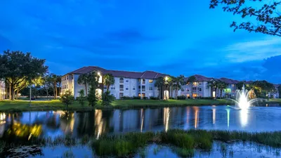 Serene lakeside apartments with elegant evening lighting and picturesque fountain views, surrounded by lush greenery under a vibrant blue sky.