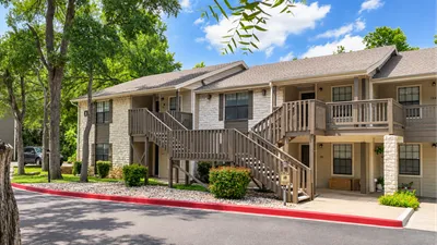 Charming apartment complex with stone accents and wooden balconies, nestled among lush greenery and bathed in sunlight.