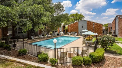 Inviting apartment complex with a sparkling outdoor swimming pool, surrounded by lush greenery and cozy seating areas under a clear blue sky.