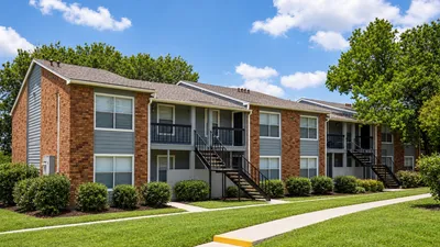Charming apartment complex with brick and siding exterior, lush green lawns, and inviting pathways surrounded by mature trees.