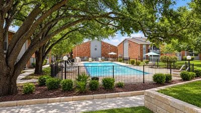 Serene apartment complex with a sparkling pool, shaded by lush trees and inviting outdoor seating.