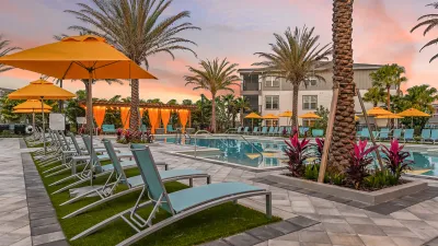 Resort-style pool area at sunset with lounge chairs, orange umbrellas, palm trees, and a covered cabana, set within a landscaped apartment community.