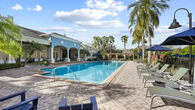 Resort-style swimming pool surrounded by lounge chairs, blue umbrellas, palm trees, and a spacious sundeck.