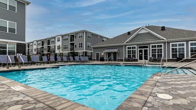 Modern apartment community featuring a sparkling outdoor pool surrounded by stylish lounge chairs, with contemporary buildings in the background under a clear blue sky.