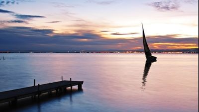 Serene lakeside view at sunset with a sailboat gliding on calm waters, creating a tranquil and picturesque atmosphere.