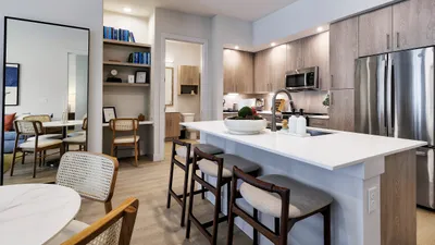 Modern apartment kitchen with sleek white countertops, stainless steel appliances, and cozy seating area. Elegant dining nook adjacent with stylish wooden chairs and decorative shelving.