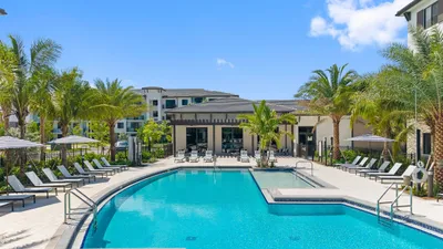 Luxurious apartment complex showcasing a sparkling outdoor pool surrounded by palm trees and elegant lounge chairs under a clear blue sky.