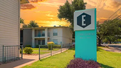 Charming apartment complex with lush landscaping and a vibrant sunset sky, highlighted by a modern teal sign at the entrance.