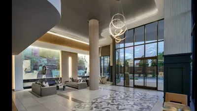 Elegant apartment lobby featuring contemporary artwork, chic seating, and a grand chandelier, framed by floor-to-ceiling windows overlooking a vibrant landscape.