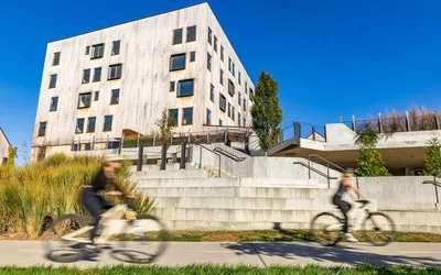 Modern apartment complex with a sleek wooden facade and landscaped surroundings, featuring cyclists enjoying the outdoor space on a sunny day.