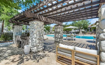 Charming outdoor space featuring a stone-pillared pergola, modern grill, and inviting pool area with sunlit seating.