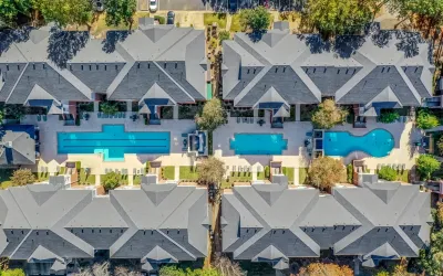 Aerial view of a modern apartment community featuring multiple resort-style swimming pools, spacious sundecks, landscaped courtyards, and contemporary residential buildings surrounded by greenery.