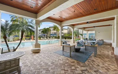 Covered poolside lounge featuring comfortable outdoor seating, wood-paneled ceilings, and views of the resort-style swimming pool surrounded by palm trees.