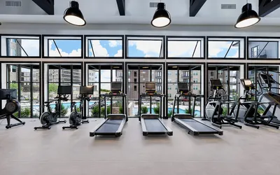 Modern apartment fitness center with state-of-the-art treadmills and exercise bikes against a backdrop of large, sunlit windows overlooking a beautiful pool area.
