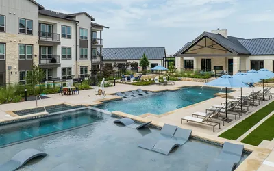 Luxurious apartment complex with a stunning outdoor pool, elegant lounge chairs under blue umbrellas, and modern architecture set against a clear sky.