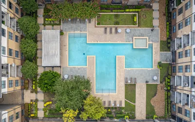 Aerial view of a luxurious apartment complex featuring a large T-shaped pool surrounded by lush greenery, modern lounge chairs, and pristine landscaping.