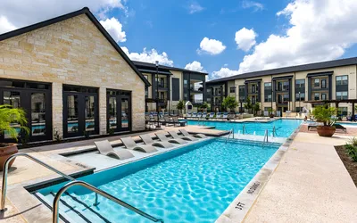 Luxurious apartment complex with a stunning outdoor pool area, featuring modern lounge chairs and a vibrant, clear blue sky.