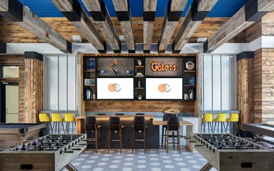 Trendy entertainment room with rustic wood accents, vibrant blue ceiling, foosball tables, plush bar seating, and dual screens perfect for gatherings at Campus Circle Gainesville apartment community.