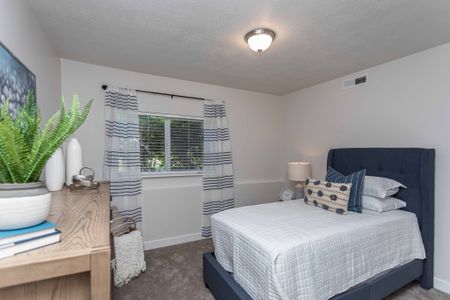 Bedroom 1 features new plush creme carpet, two tone paint, and a large window for natural light. There is a blue upholstered twin sized bed with a white comforter and coordnating throw pollows. Room shows it can comfortably accomidate a che