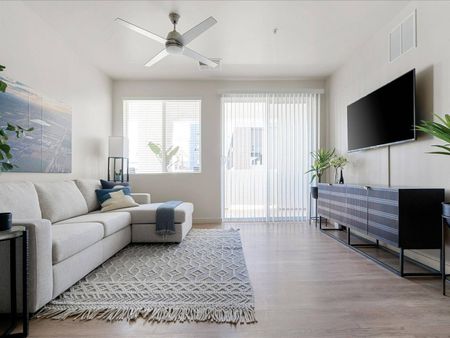 One, Two, & Three-BR Apartments In Glendale, AZ - Acero At The Stadium - Living Room With Sectional, Area Rug, End Tables, Cabinet, TV, Sliding Glass Door, Ceiling Fan, And Window.