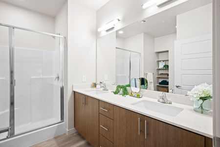 Goodyear AZ Apartments - Acero Roosevelt - A Bright And Spacious Bathroom With Wood-Style Flooring, Dark Cabinets, A Shower, And A Two Sink Vanity With A Huge Mirror And White Countertops