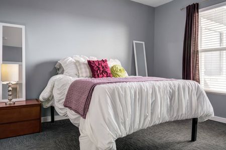Model apartment bedroom with gray patterned carpet, gray walls, and furniture including a full-size bed and nightstands.