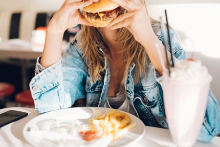Stock photograph of someone sitting down and eating a large burger. There are fries and a milkshake on the table.