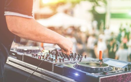 Stock photo of a DJ using a mixing board. The photo has a colorful filter and there is a crowd in the background.