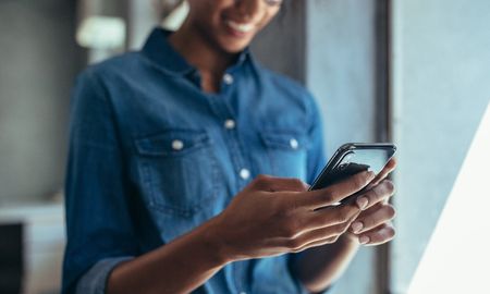 Stock photograph of somebody using their phone. They are wearing denim, glasses, and smiling widely.