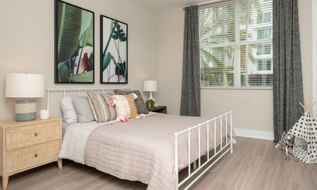Model bedroom at our apartments in Pompano Beach, FL, featuring wood laminate floors, large windows, and pink bedspread.