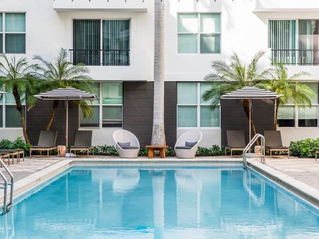 The pool area at our apartments for rent in Pompano Beach, featuring umbrellas, beach chairs, and palm trees.