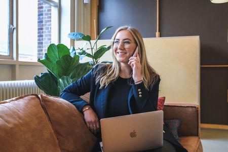 woman sitting on leather couch talking on cell phone with laptop