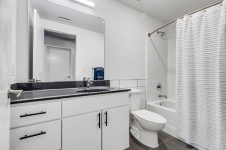 A contemporary bathroom featuring a wide vanity with a dark granite countertop and a white undermount sink. Above the sink, there's a large mirror framed by wall sconces on either side. A white toilet is positioned next to a bathtub, which is concealed by a white, textured shower curtain.