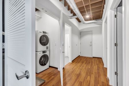 : A well-lit entry hallway that leads deeper into the home. To one side, a modern laundry room is visible, boasting a stacked white washer and dryer set. The floor is adorned with rich wooden planks, and the white walls are complemented by exposed overhead ductwork and wooden beams, giving the space a blend of contemporary and industrial vibes. The hall is further defined by white louvered closet doors and a clear path guiding towards other parts of the residence.