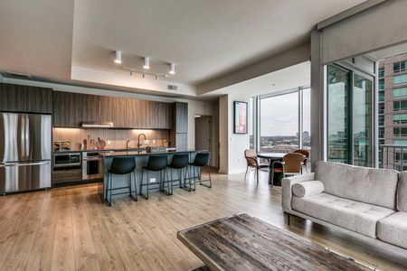 Wide view of open concept apartment home with kitchen with stainless steel appliances and rich lighting, kitchen island, and dining area, with downtown views in the background
