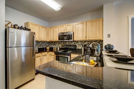 U-shaped kitchen with black countertops, stainless steel refrigerator, microwave and stove. Ample cabinet space with blonde cabinets above and below counter with multi-colored tile backsplash