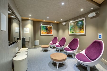 Side view of game room with oversized TV built into wall. Four, small and white, round stools are placed in a row below TV with four purple and white bucket chairs to the right. Two wood and white ottomans are placed in front of bucket chai