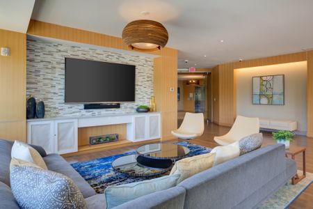 View of stylishly designed resident lounge from behind sectional sofa. Mirrored nesting coffee table sits in front of sectional on multicolored blue rug facing large wall mounted TV with tile wall behind. Two white bucket chairs are placed