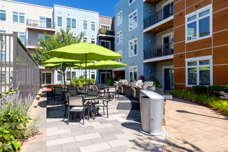 Outdoor space with four top tables and chairs; lime green umbrellas overhead. Three gas grills and silver trash receptacle with apartment building in background.