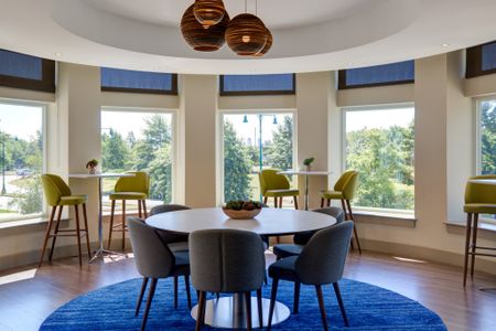 Resident lounge with round, six person table and chairs placed on round bright blue rug with round lantern lights hung above. Three high top tables with lime green stools are placed in front of windows in circular space.