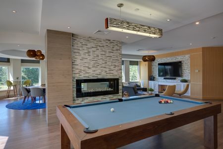 View of resident lounge behind pool table with blue felt top with fireplace in front. Large wall mounted TV and seating can be seen to the right and tables and chairs with large windows are to the left.