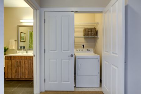 Bathroom vanity with wood patterned cabinets below vanity and silver framed mirror hung above seen to left. Laundry room with one door closed and one door open showing white dryer and two shelves for storage above is on the right.